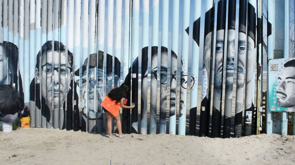 Dr. Lizbeth De La Cruz Santana paints one of the faces on the border wall at the Playas de Tijuana Mural Project