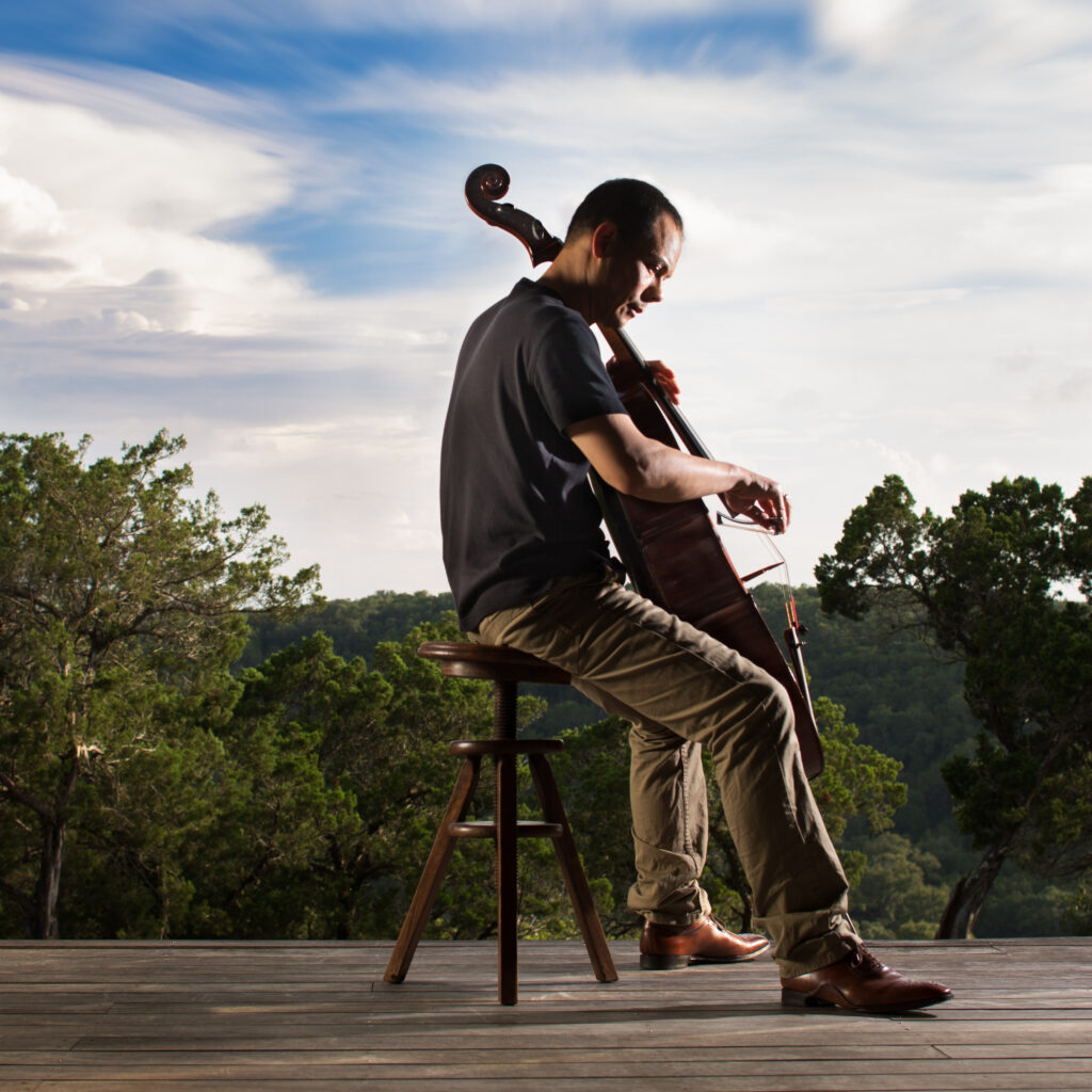 Bion Tsang plays his cello on a wooden deck surrounded by trees.