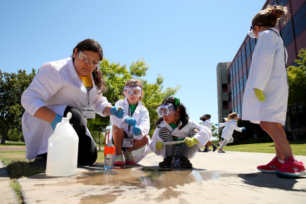 A Fresno State student helps elementary aged students, wearing goggles, with a wet science experiment.