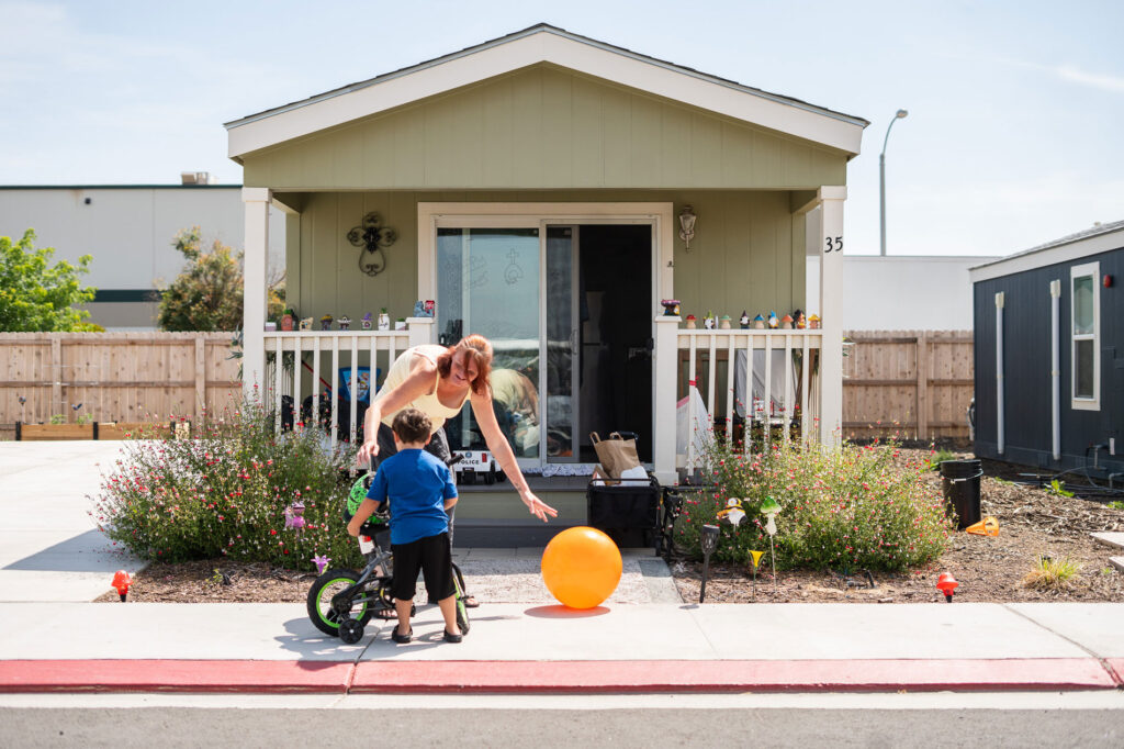 A woman and child with a bicycle and large orange ball on the sidewalk in front of a small home.