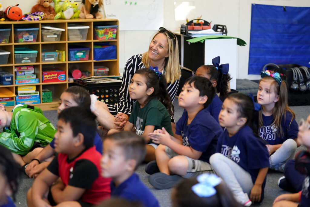 Dr. Corrine Folmer sits with elementary age kids on a mat and smiles.