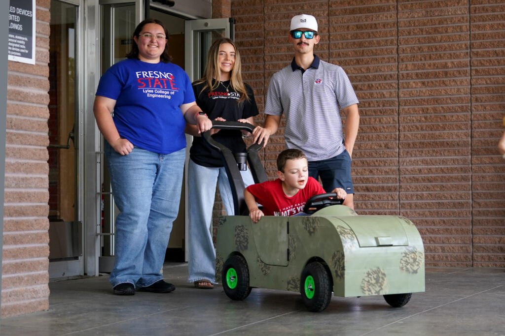 A young boy drives a custom-built camouflage ride-on toy car while three Fresno State engineering students stand behind him smiling.