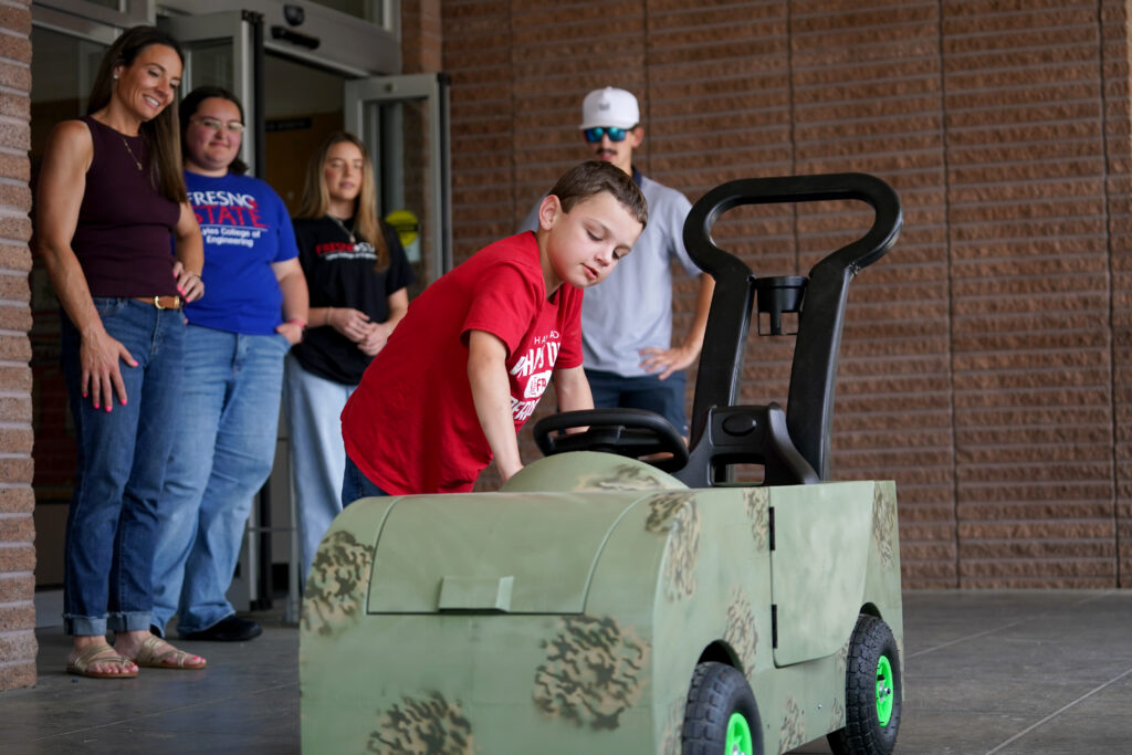 A young boy examines his new custom-built toy car as students and family members look on.