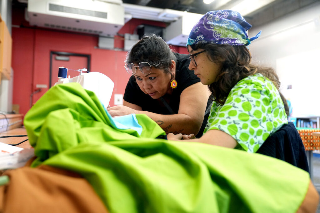 A student and a woman look closely at a sewing machine while making a shawl.